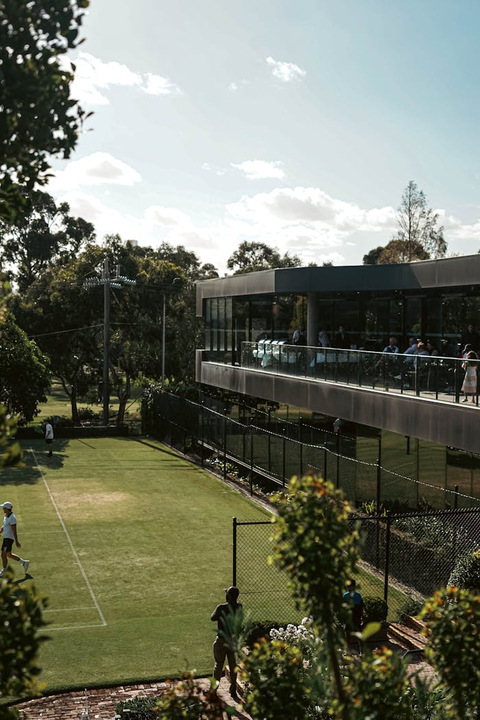 gallery-4 Beautiful outdoor tennis court next to a modern clubhouse under a clear blue sky.