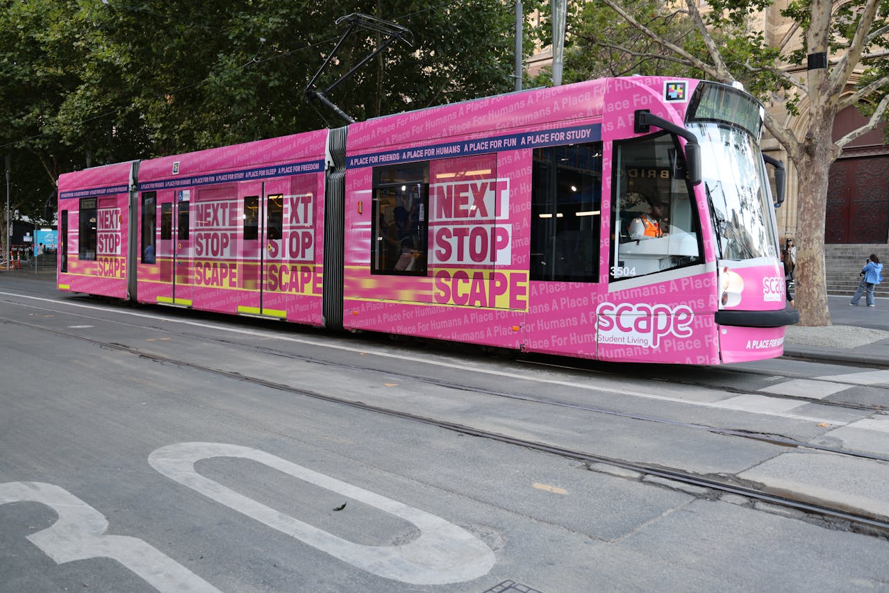 services-01 Bright pink tram on a city street with 'Next Stop Scape' advertisement, capturing urban life.